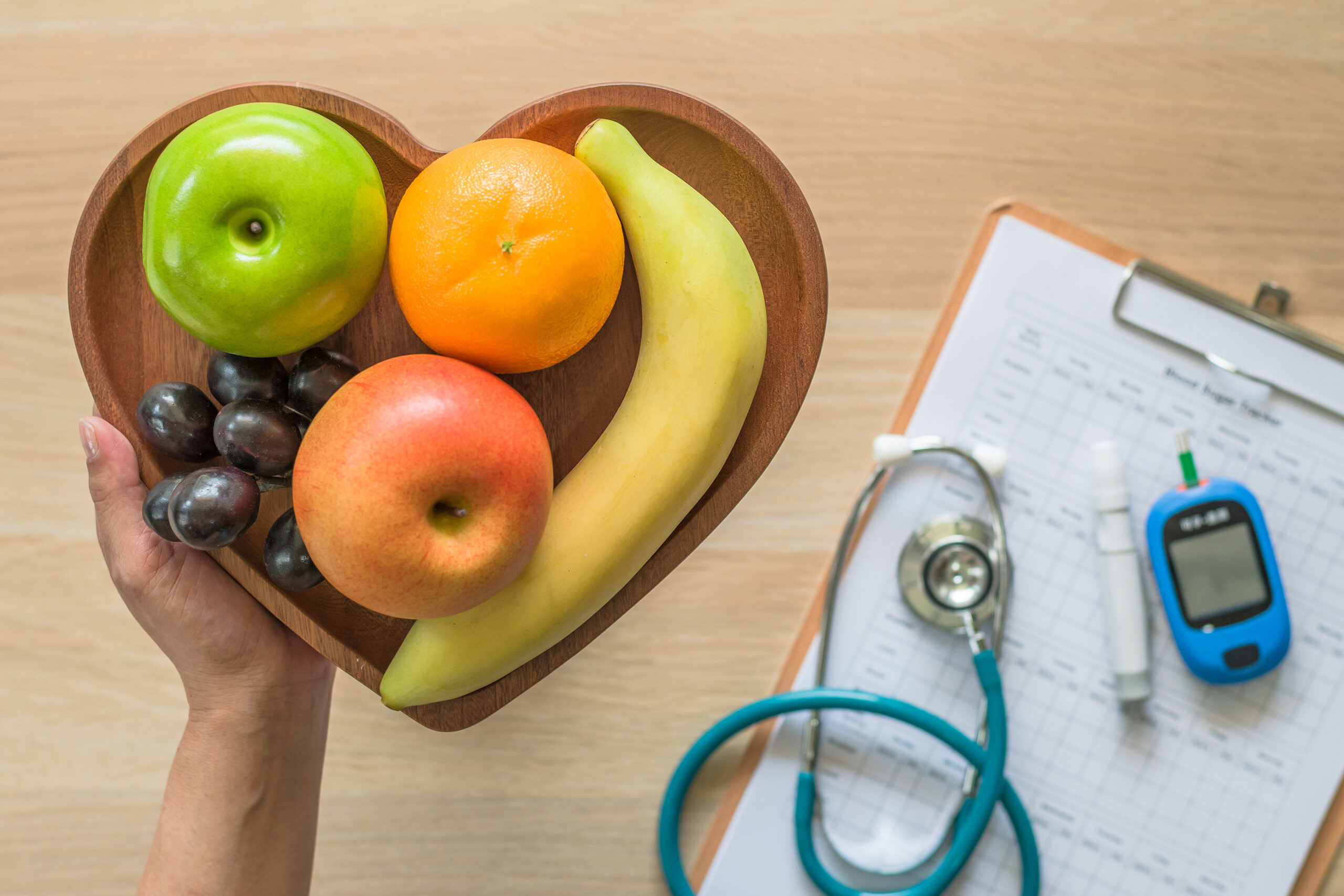 A person holding a heart-shaped wooden bowl filled with colorful fruits including green and red apples, an orange, grapes, and a banana, with a stethoscope and glucose meter in the background.